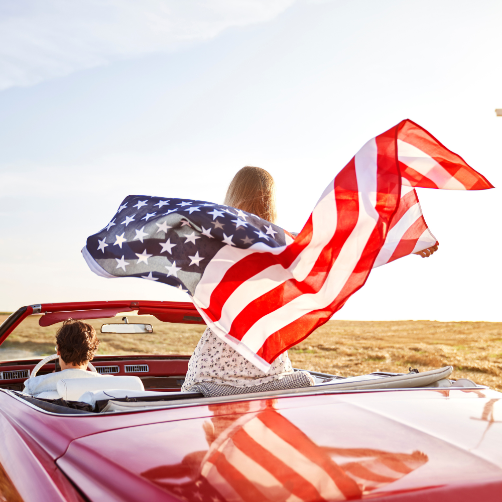 A person waves a US flag out of a window of a convertible car on Route 66